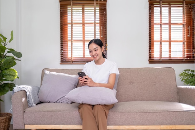 Happy young beautiful woman is playing social media on her phone while sitting on the sofa at home