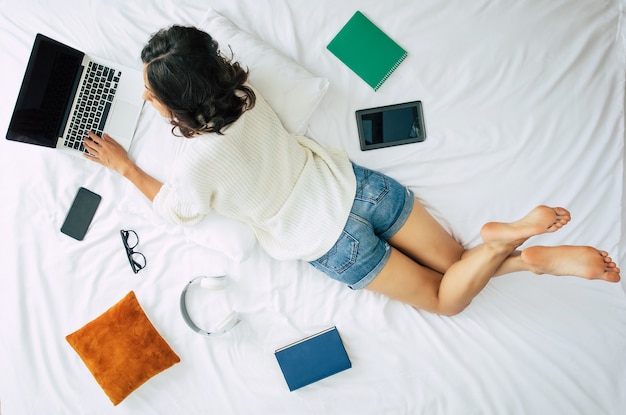 Happy young beautiful woman is working on the laptop while lying on bed at home. Top view or above photo