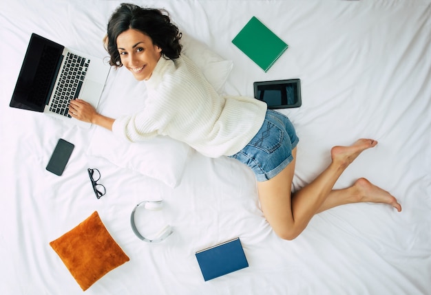 Happy young beautiful woman is working on the laptop while lying on bed at home. Top view or above photo