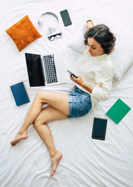Happy young beautiful woman is working on the laptop while lying on bed at home. Top view or above photo