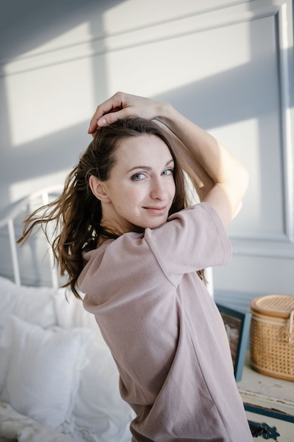 Happy young beautiful woman pulls her hands up, standing next to the bed in her bedroom at home. People and Morning concept.