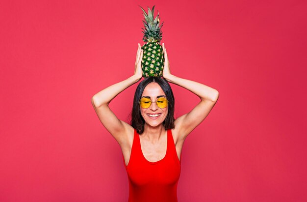 Happy young beautiful woman in red swimsuit sunglasses and big summer hat with pineapple in sunglasses in hand posing over pink background