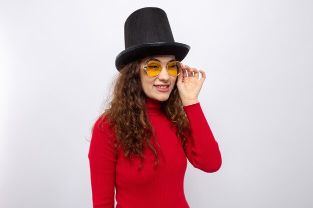 Felice giovane bella donna in dolcevita rosso con cappello a cilindro che indossa occhiali gialli che guarda da parte sorridendo allegramente in piedi su bianco Foto Premium