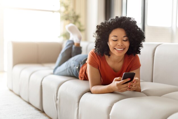 Happy young black female using smartphone while lying on comfy sofa at home