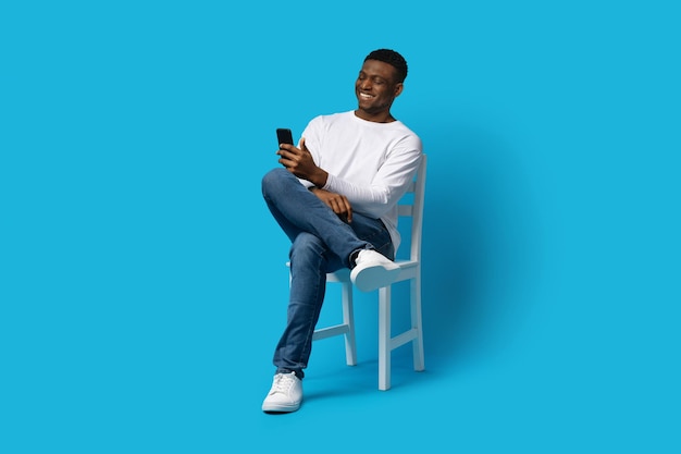 Happy young black man sitting on chair using cell phone