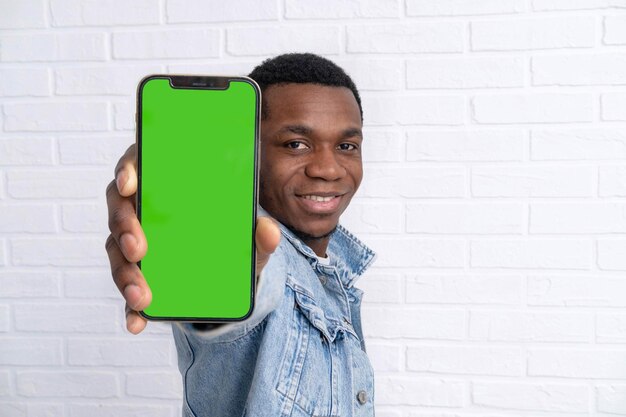 Happy young black man standing and pointing Big smartphone with blank green screen demonstrating copy space for app or website design standing over white brick background mockup image