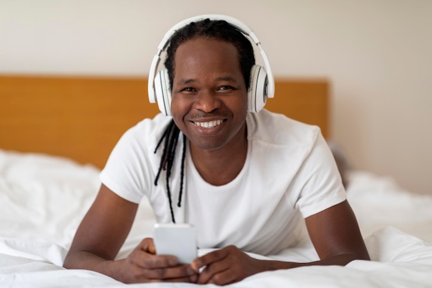 Happy Young Black Man Wearing Wireless Headphones Relaxing In Bed With Smartphone