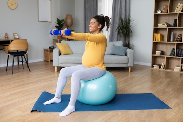 Happy young black pregnant woman with big belly doing exercises on fitness ball with dumbbells