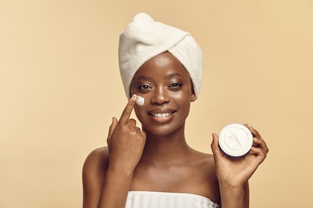 Happy young black skin woman holding jar of cream wearing in\
spa towel and head turban over beige background eco friendly\
cosmetics and beauty products for body and face care concept