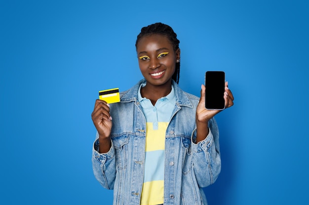 Happy young black woman showing bank card and smartphone mockup