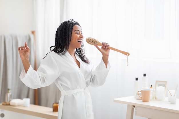 Happy young black woman singing in bathroom using body brush as microphone