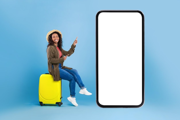 Happy young black woman sitting on suitcase and pointing at smartphone with mockup for online travel