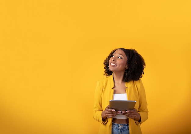 Happy young black woman using digital tablet on yellow studio background looking at copy space and