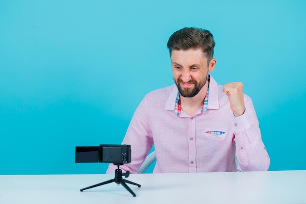 Happy young blogger man is raising up his fist by sitting in front of his mini camera on blue background