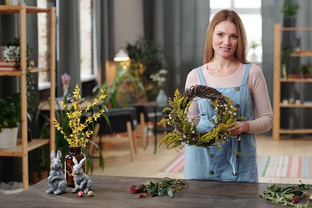 Happy young blond female in casualwear looking at you with smile while standing by wooden table in home environment and showing wreath