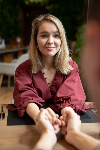 Happy young blond female in elegant dress looking at her boyfriend while holding his hands by served table in modern restaurant or cafe