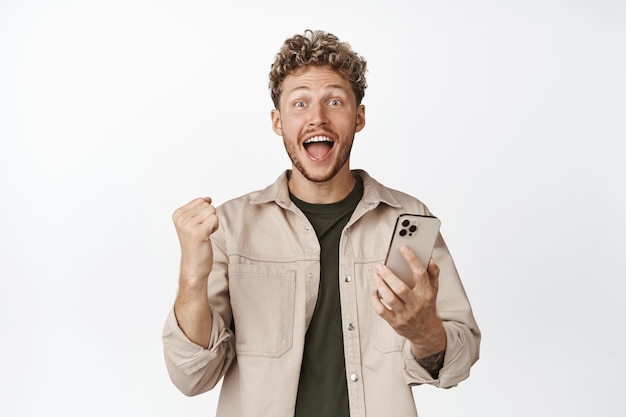 Photo happy young blond man holding mobile phone and screaming joyful winning on smartphone celebrating achievement on cellphone standing against white background