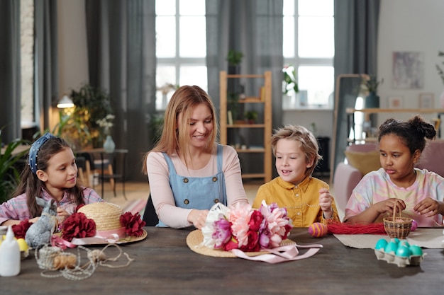 Happy young blond woman helping cute little children with decorating hats with handmade flowers of vivid colors while sitting by table