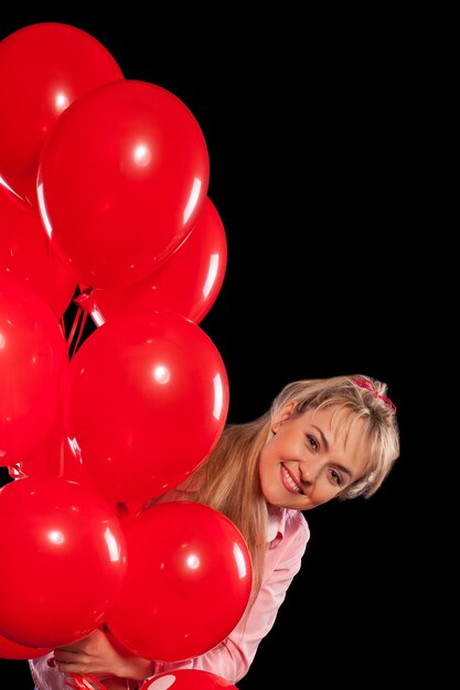 Happy young blond woman looking out of red balloons and smiling. Isolated on black background