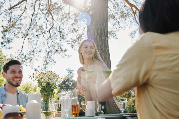 Счастливая молодая блондинка берет пластиковый контейнер с салатом за сервированным столом во время ужина со своими друзьями под сосной