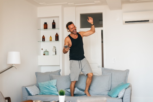 Happy young blonde man standing in the couch at home on the mobile phone listening music and dancing