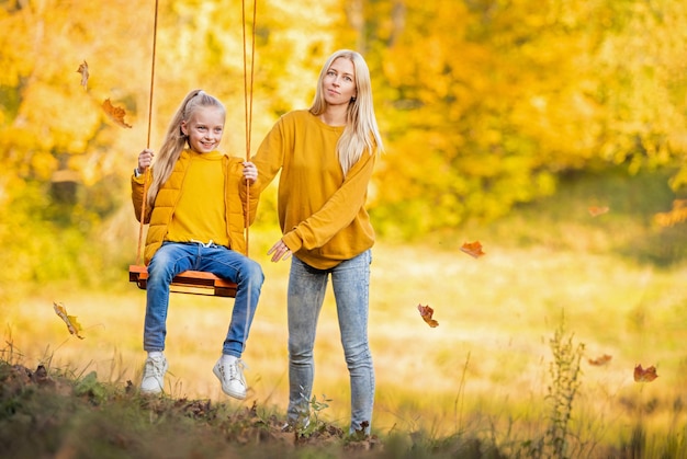 Happy young blonde mom with loose hair shakes her daughter on a swing on a rope swing in autumn in the park