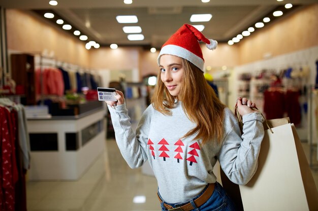 クレジットカードとカラフルな買い物袋を持って店でカメラを見てクリスマスの帽子をかぶった幸せな若いブロンドの女性新年の休日のセールでモールで女の子のショッピングギフト