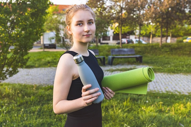 La giovane donna bionda felice tiene il tappetino da yoga e la bottiglia d'acqua nel parco fitness sportivo per uno stile di vita sano