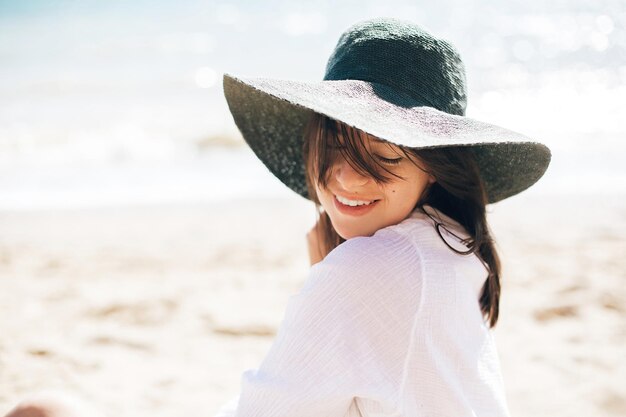 Happy young boho woman in hat relaxing and enjoying sunny warm day at ocean Space for text Stylish hipster girl sitting on beach and tanning Summer vacation Calm emotions