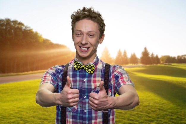 Photo happy young boy is showing thumbs up. smiling schoolboy dressed in checkered shirt with bowtie. sunny green valley on the background.