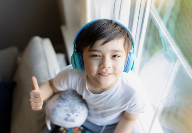 Happy young boy wearing headphones listening to music Cute kid looking up to camera with smiling face Positive Child sitting next to window relaxing on weekend in Spring or Summertime