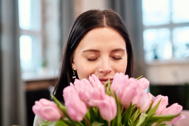 Happy young brunette female keeping her eyes closed while holding bunch of fresh pink tulips by her nose and enjoying their fragrance