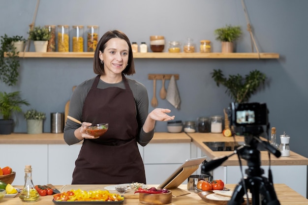 Happy young brunette woman in apron standing by kitchen table