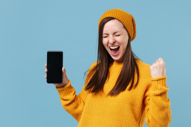 Happy young brunette woman girl in yellow sweater hat posing isolated on blue background. People lifestyle concept. Mock up copy space. Hold mobile phone with blank empty screen doing winner gesture.