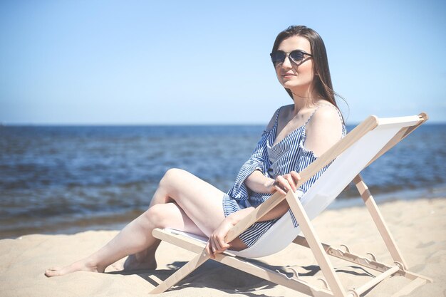 幸せな若いブルネットの女性は、笑顔で、ファッション サングラスをかけながら、海のビーチの木のデッキ椅子でリラックスします。楽しむ休暇のコンセプト