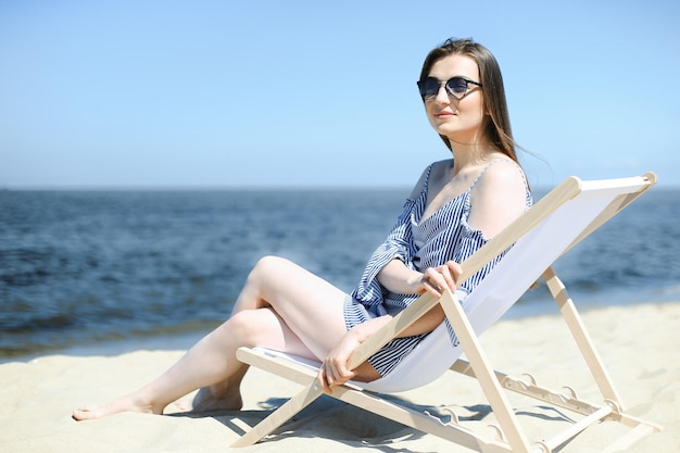 Счастливая молодая брюнетка отдыхает на деревянном шезлонге на берегу океана, улыбаясь и в модных солнцезащитных очках. Концепция отдыха