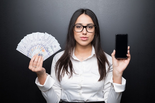 Happy young brunette woman in white shirt showing smartphone with blank screen and cash money in hands isolated on black wall