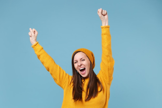 Happy young brunette woman in yellow sweater and hat posing\
isolated on blue background studio portrait. people sincere\
emotions lifestyle concept. mock up copy space. clenching fists\
like winner.