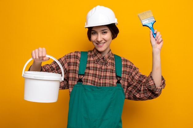 건설 유니폼을 입은 행복한 젊은 건축업자 여성, 페인트 통과 브러시를 들고 주황색 벽 위에 즐겁게 서서 웃고 있는 안전 헬멧