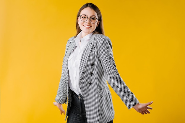 Счастливая молодая деловая женщина в куртке и рубашке в очках улыбается и позирует на камеру на желтом фоне