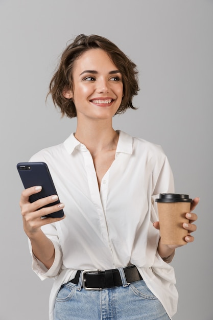 コーヒーを飲む携帯電話でおしゃべり灰色の壁の上に孤立してポーズをとって幸せな若いビジネス女性