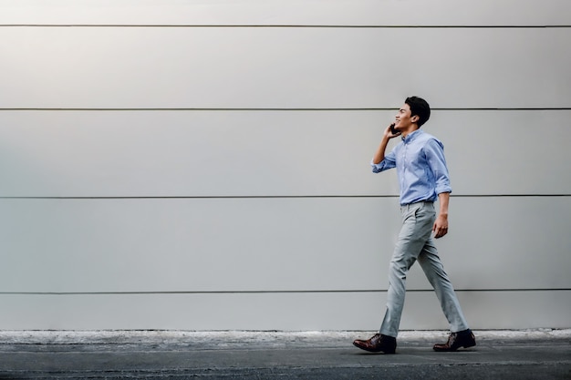도시 건물 벽으로 걷는 동안 휴대 전화를 사용 하여 캐주얼에 행복 한 젊은 사업가. 현대인의 라이프 스타일.