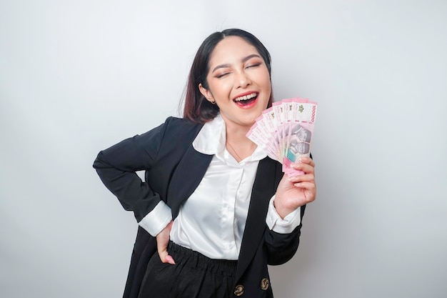 행복한 젊은 여성 사업가가 검은색 정장을 입고 흰색 배경에서 격리된 인도네시아 루피아로 현금을 들고 있습니다.