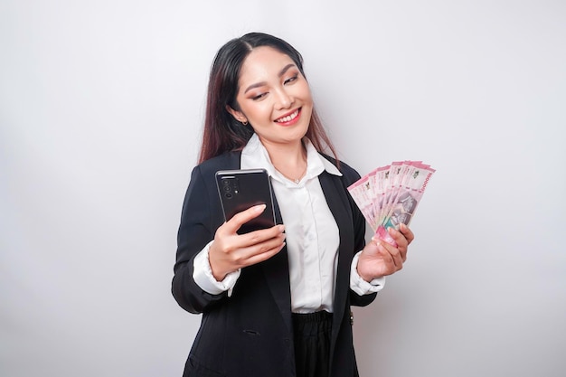 A happy young businesswoman is wearing black suit holding her phone and money in Indonesian rupiah isolated by white background