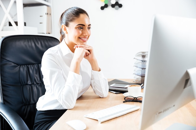 Happy young businesswoman looking at computer screen while sitting at the office table