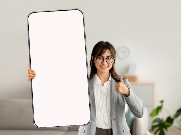 Happy young businesswoman showing thumb up gesture demonstrating cellphone with mockup for website