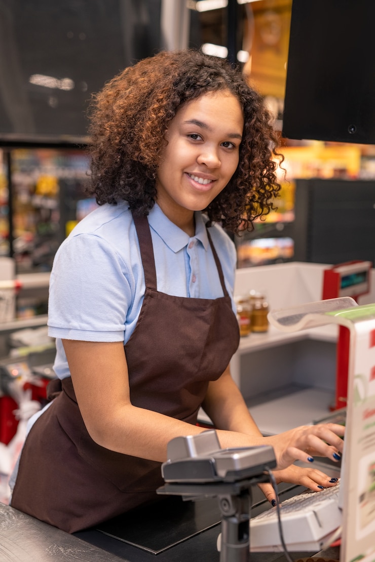 Photo happy young cashier with dark wavy hair looking at you while standing by workplace and pressing button of cashbox