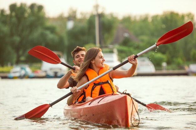 Happy young caucasian couple kayaking on river with sunset in the backgrounds. Having fun in leisure activity. Happy male and female model laughting on the kayak. Sport, relations concept. Colorful.