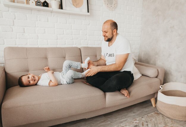 Happy young caucasian father on sofa in living room playing with little preschool son