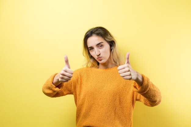 Happy young caucasian female in an orange sweater making thumb up sign and smiling